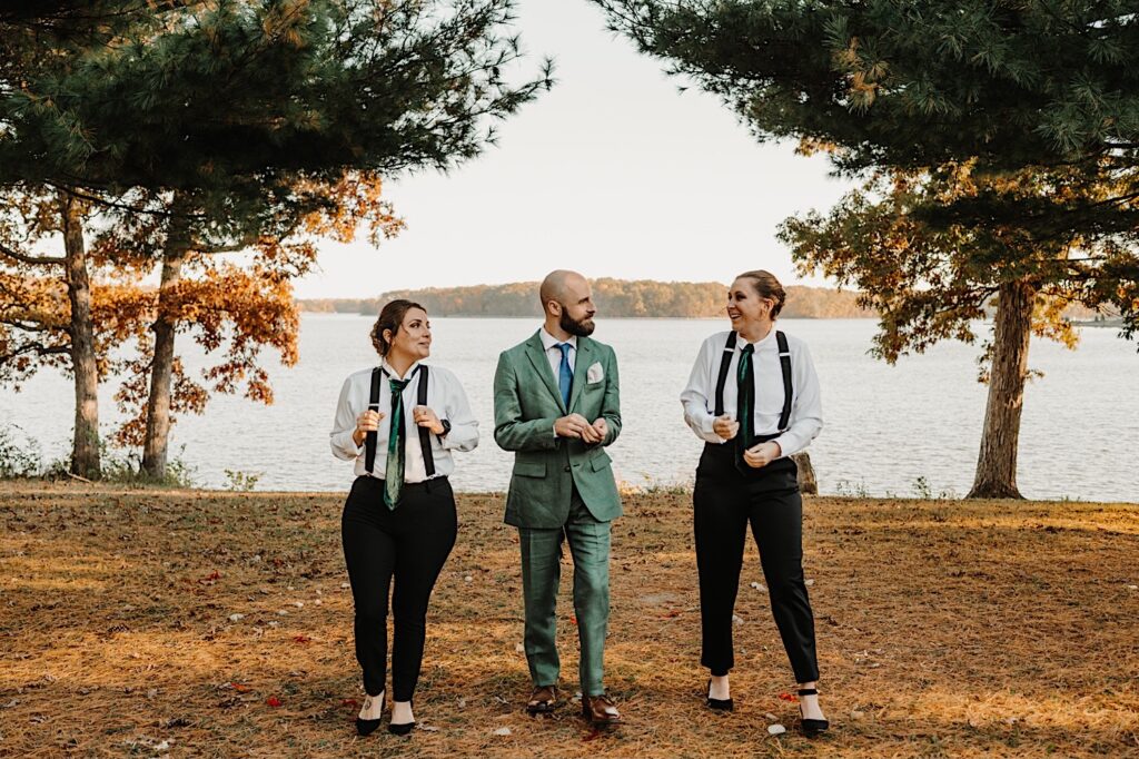A groom walks with the two members of his wedding party on either side of him towards the camera as the three joke with one another, behind them is a lake