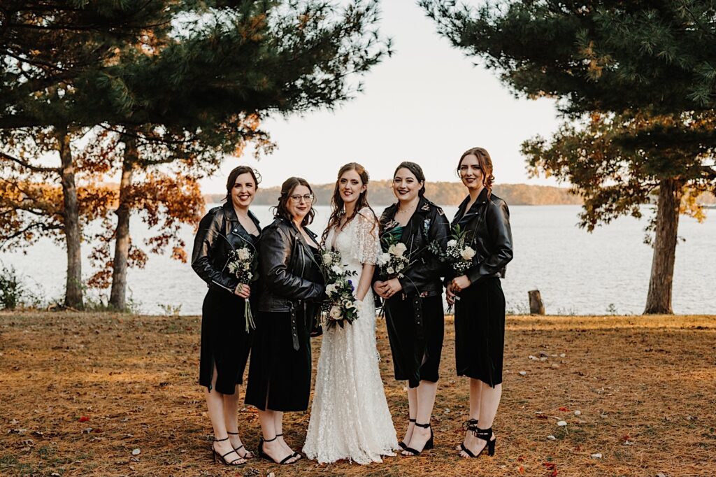 A bride stands with her 4 bridesmaids on either side of her as they all smile at the camera, behind them is a lake