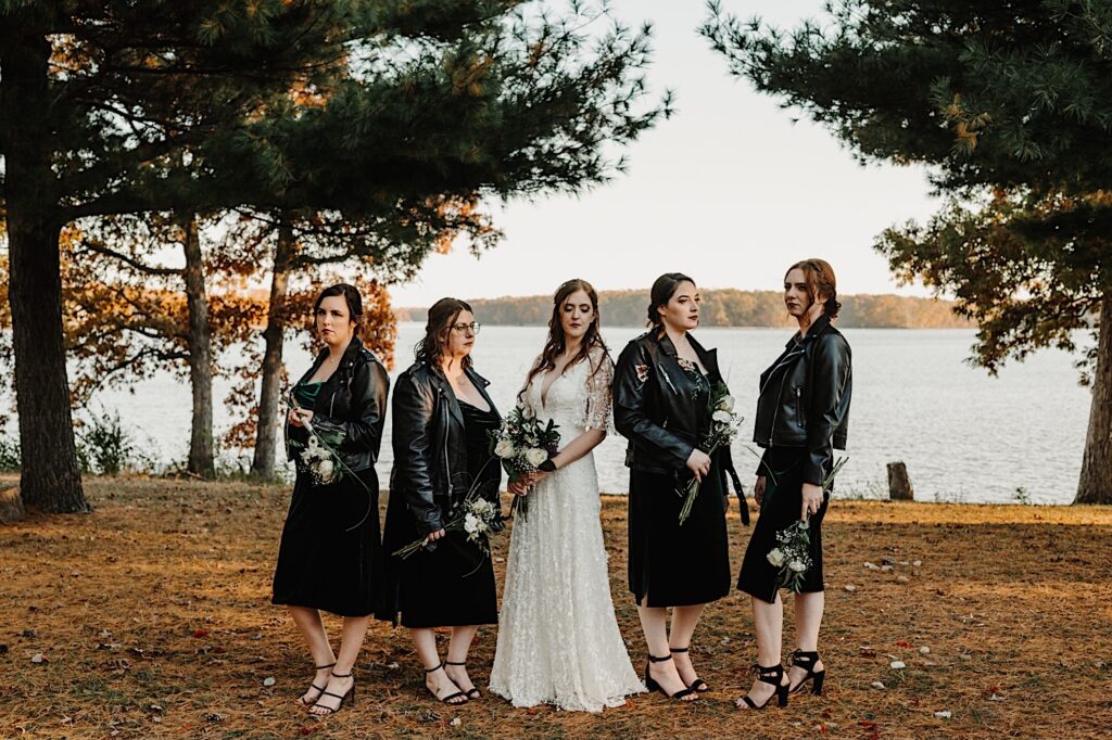 A bride stands in between her 4 bridesmaids as they all pose for a serious photo with a lake behind them