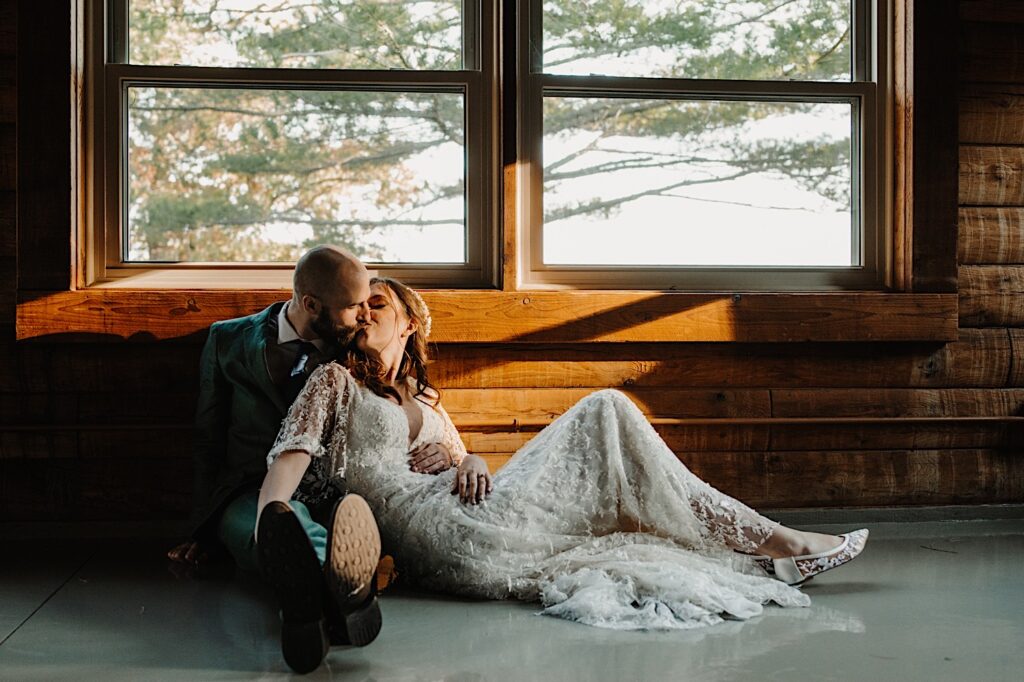 A bride and groom sit on the floor inside a log cabin and kiss one another with a window above them