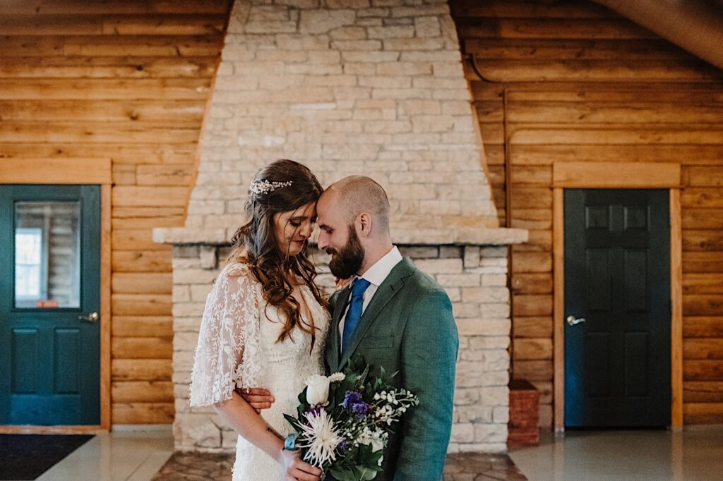 After their intimate fall wedding ceremony at a lake near Bloomington Illinoi a bride and groom stand together in front of a fire place inside a log cabin and smile at one another while touching their foreheads together
