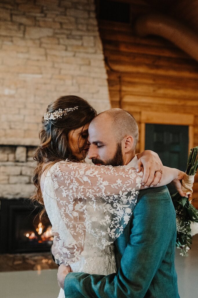 A bride and groom hug one another inside of a log cabin in front of a fireplace