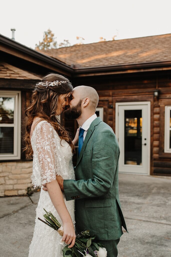 A bride and groom embrace as they are about to kiss one another while standing in front of a log cabin