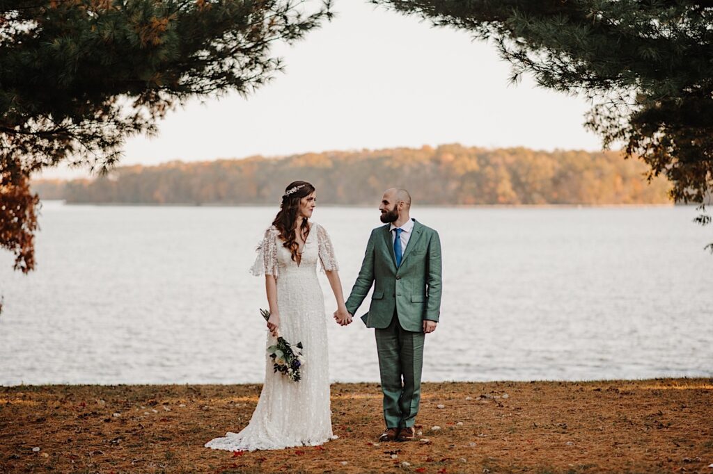 At the site of their intimate wedding ceremony space in front of a lake near Bloomington Illinois a bride and groom stand and hold hands while looking at one another