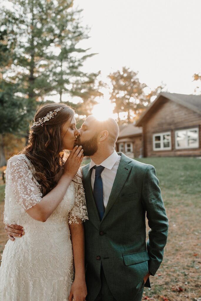 A bride and groom kiss one another while standing in front of a log cabin as the sun begins to set behind them