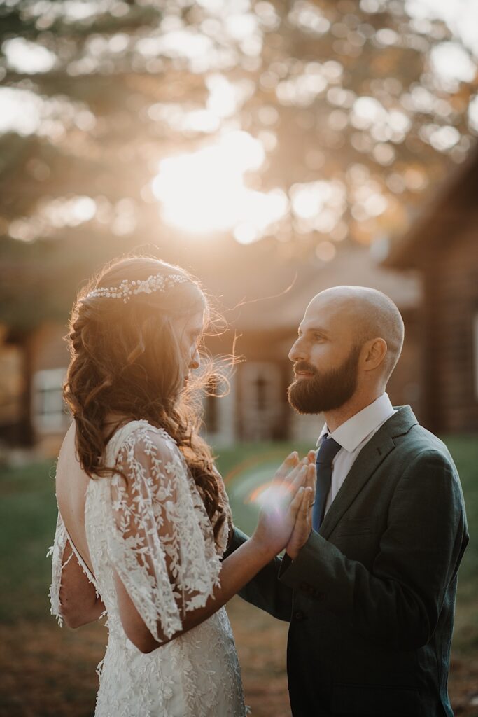 A bride and groom look at one another and touch their palms together as the sun sets on the log cabin behind them