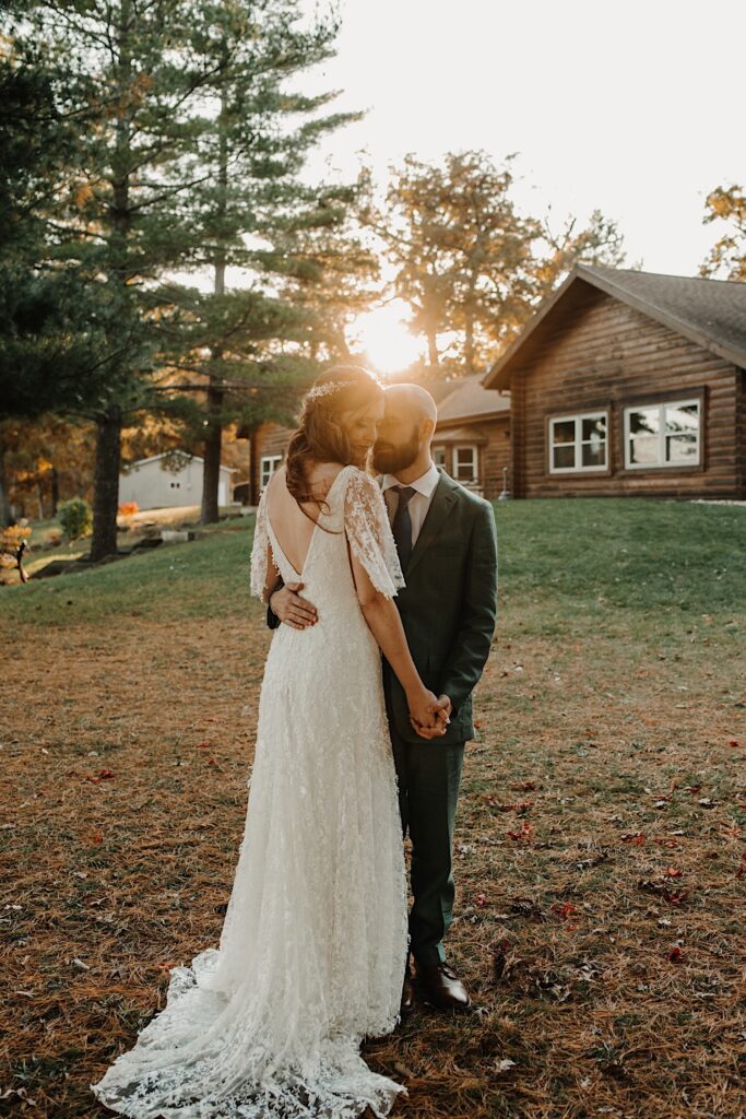 A bride faces away from the camera and embraces the groom, she is looking down over her shoulder as the groom looks at her and the sun sets on the log cabin behind them