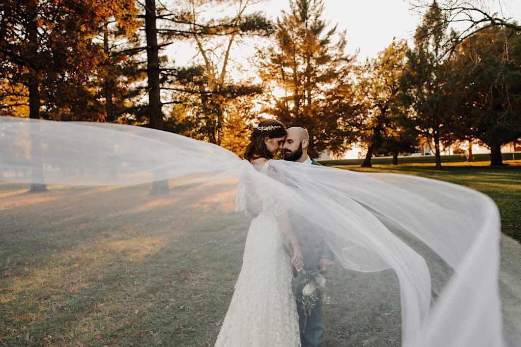 After their intimate fall wedding ceremony near Bloomington Illinois a bride and groom stand side by side with their eyes closed and the bride's veil flows towards the camera while the sun sets through the trees behind them
