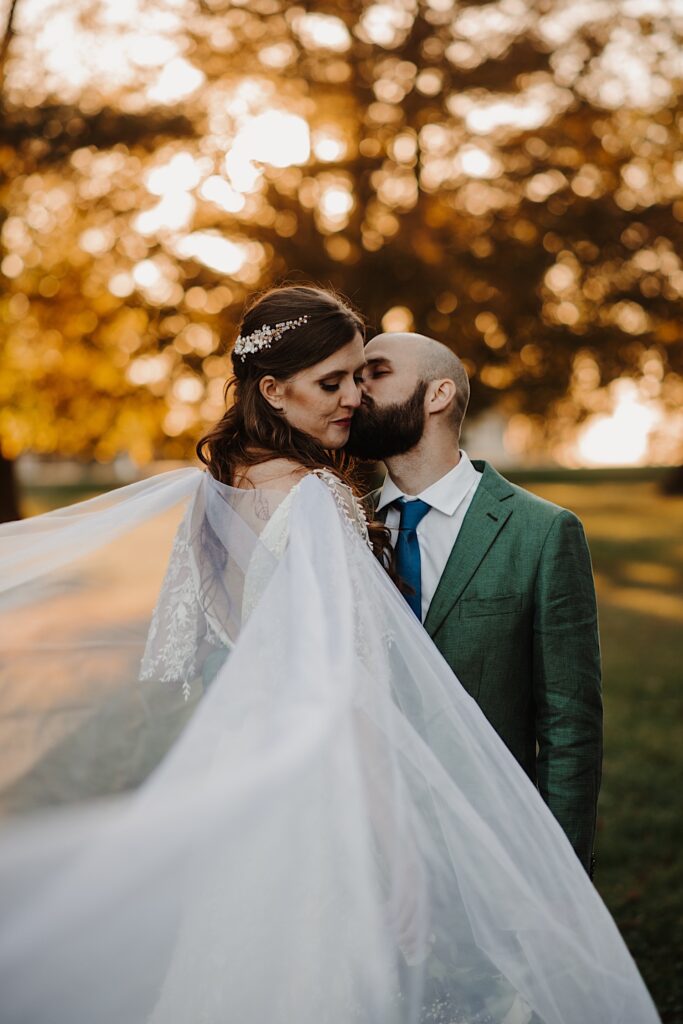 A bride smiles as the groom kisses her cheek while they face one another and her veil flows towards the camera while the sun sets through the trees behind them