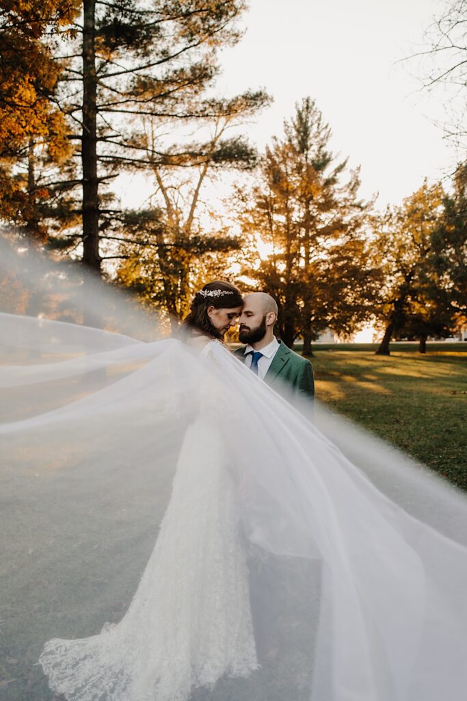 A bride and groom touch foreheads together with their eyes closed while the sun sets on the trees behind them and the bride's veil flows towards the camera