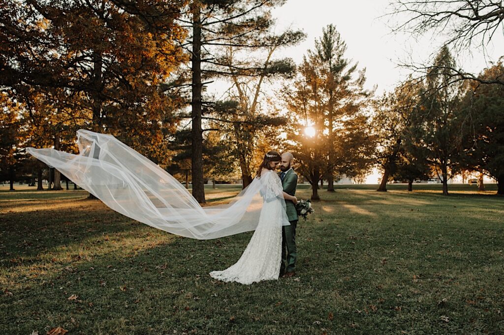 After their intimate fall wedding ceremony near Bloomington Illinois a bride and groom stand in a field surrounded by trees and embrace while the sun sets and the bride's veil flows in the wind behind her
