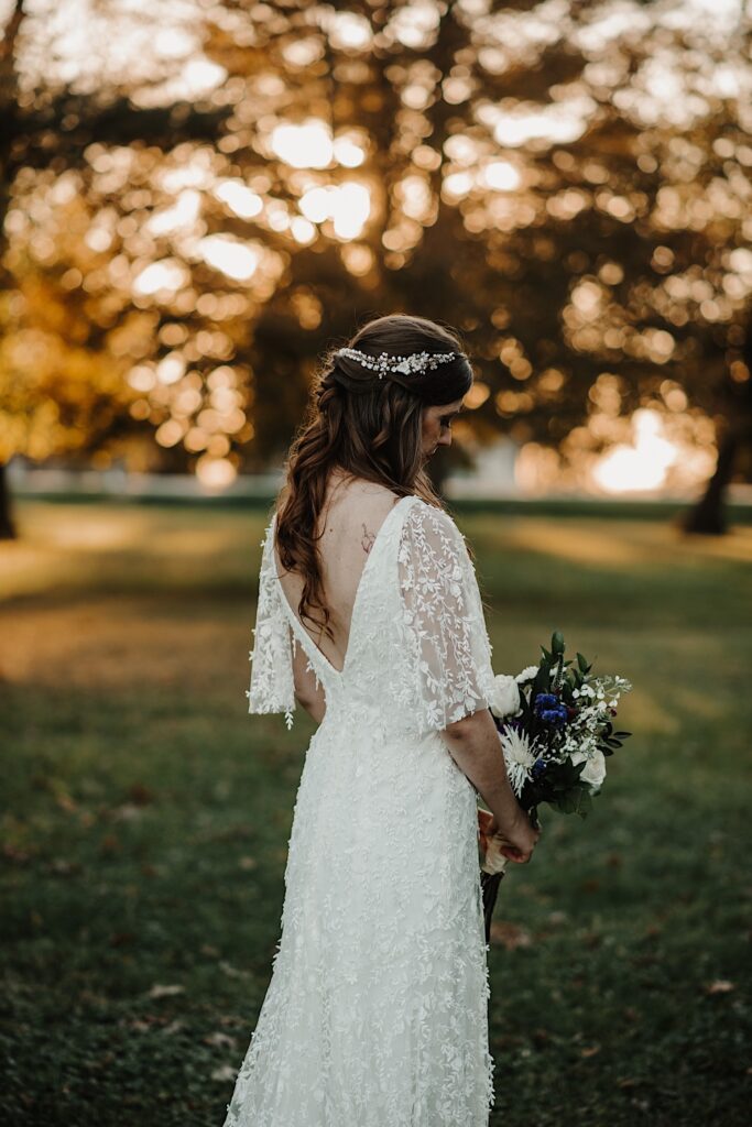 Portrait photo of a bride standing facing away from the camera and looking down at her bouquet while the sun sets on the trees behind her