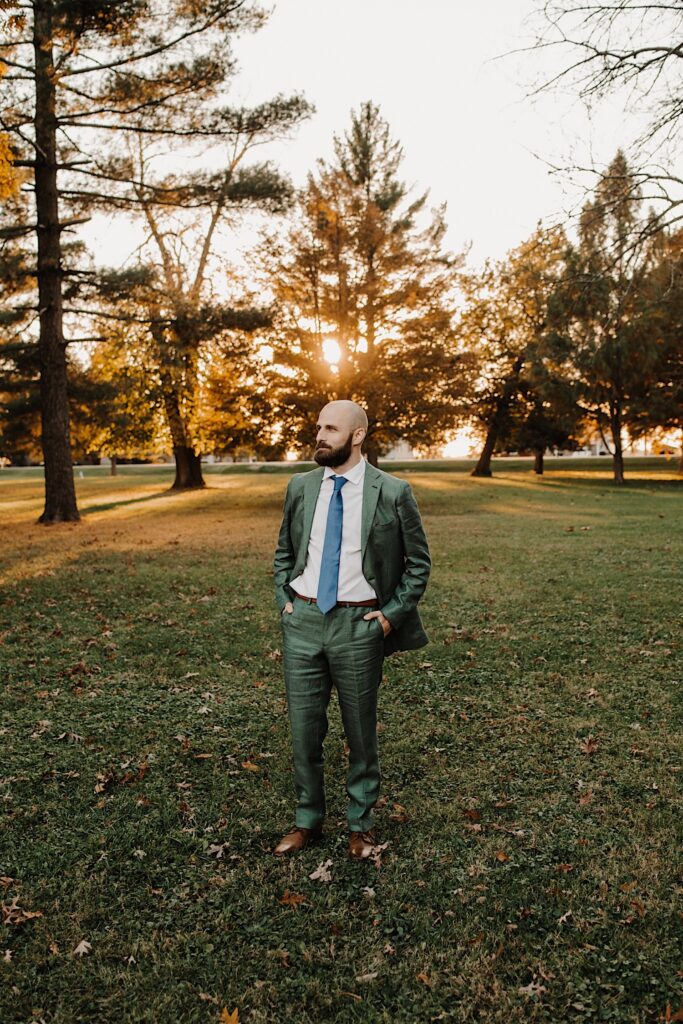 Portrait photo of a groom standing in a field surrounded by trees at sunset with his hands in his pockets looking to the left