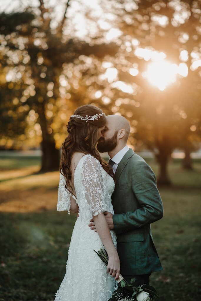 A bride and a groom are about to kiss one another while surrounded by trees as the sun sets behind them