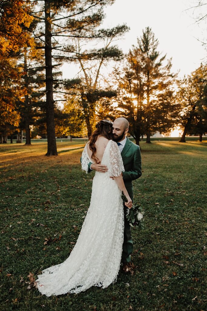 A bride facing away from the camera is hugged by the groom while the two stand in a field surrounded by trees as the sun sets
