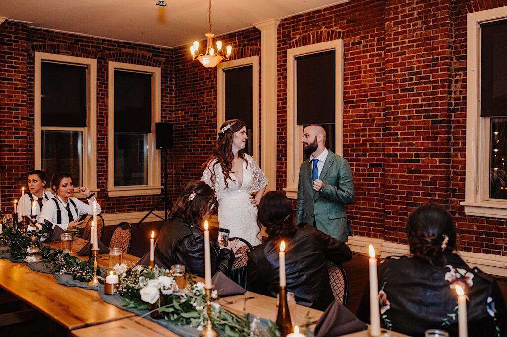 A bride and groom stand next to one another and smile at each other in front of a brick wall while their wedding parties are seated at the table in front of them during their intimate indoor wedding reception in the fall in Bloomington Illinois