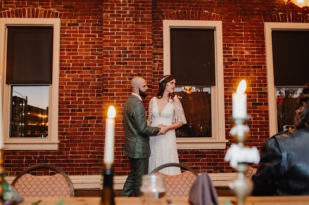 During their intimate indoor fall wedding reception at their wedding venue in Bloomington Illinois a bride and groom hold hands while looking at something to the right off camera during their first dance