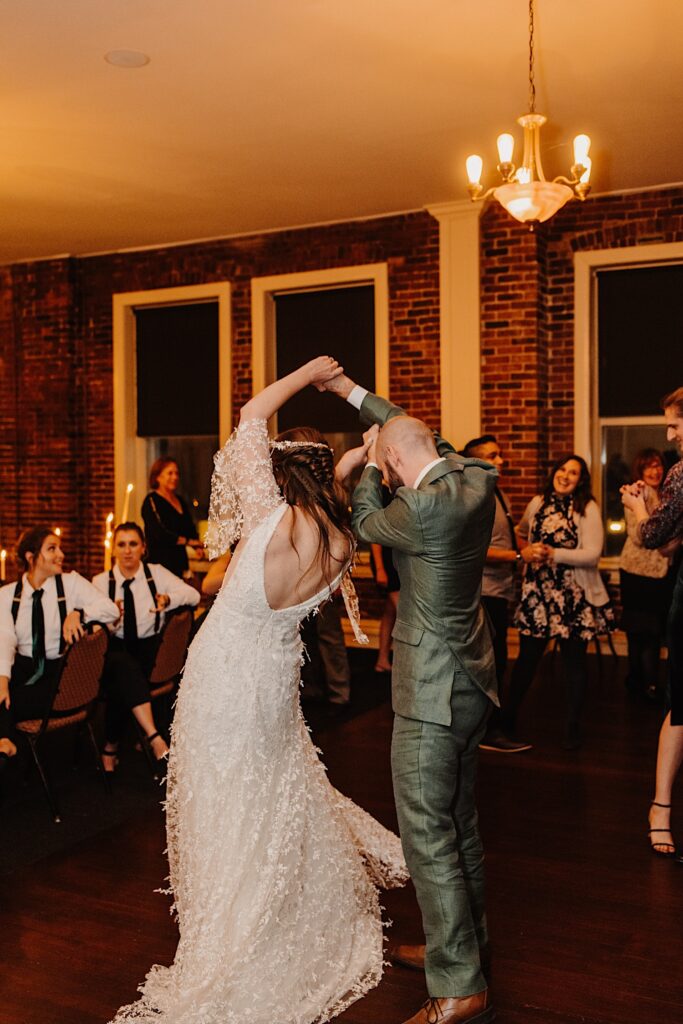 A bride is spun by the groom while they dance at their indoor wedding reception as guests behind them either sit or dance
