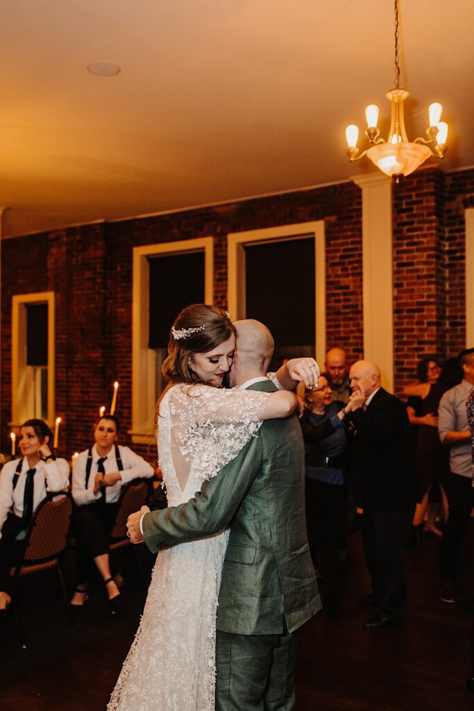 A bride smiles as she hugs the groom during their first dance at their indoor wedding reception, guests can be seen behind them either seated or dancing as well