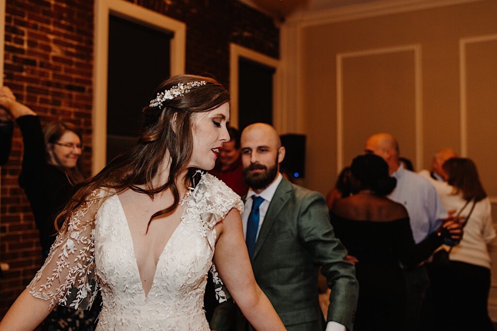 During an indoor intimate fall wedding reception at a venue in Bloomington Illinois a bride looks over her shoulder to the groom who is standing behind her as the two dance surrounded by other dancing guests of the wedding