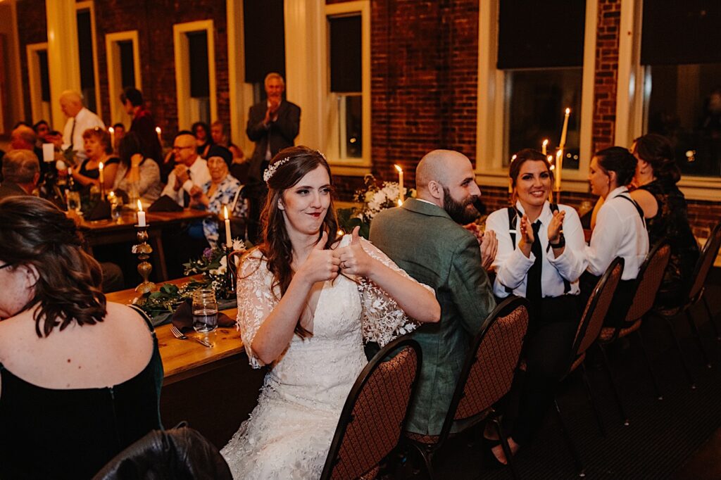 A bride sitting next to the groom gives two thumbs up while the groom and the wedding party members seated next to them clap at something off camera, this is taking place inside their Bloomington Illinois wedding venue during their intimate fall wedding reception