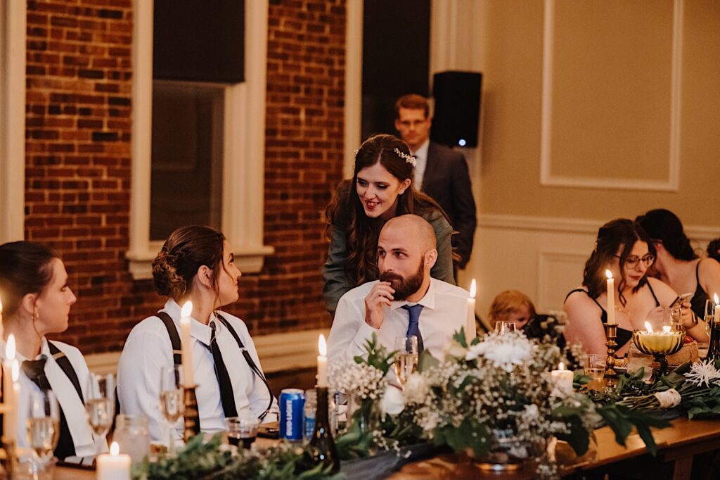 A bride smiles while standing behind the groom who is sitting and looking at members of his wedding party who are also sitting next to them during their intimate fall wedding reception at their indoor venue in Bloomington Illinois