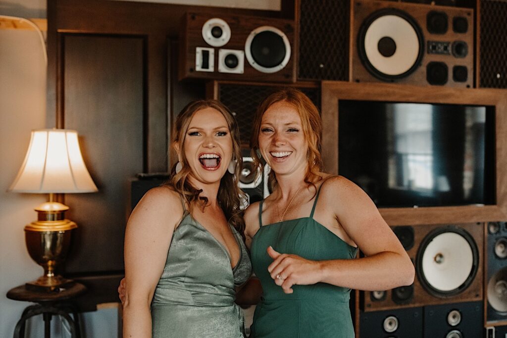 Two bridesmaids pose next to one another and smile at the camera during a wedding at The Ivy House in Milwaukee, they're in a getting ready space that has a ton of speakers and a tv on the wall behind them