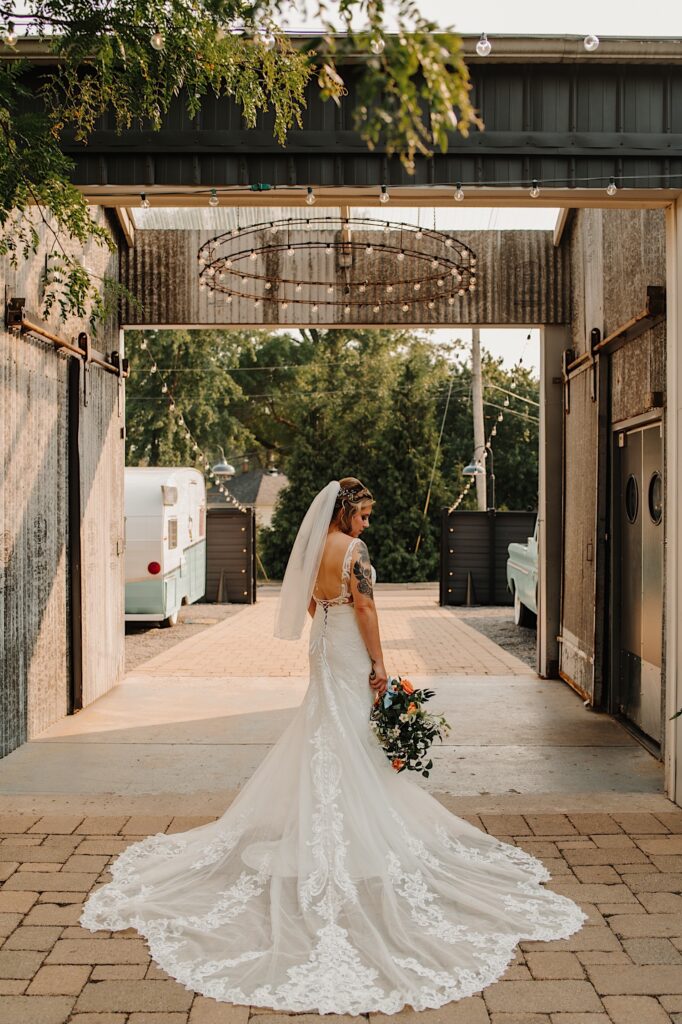 A bride stands facing away from the camera in an outdoor courtyard with a chandelier overhead and looks down at her bouquet