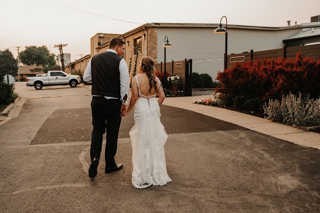 A bride and groom walk along a street in Chicago towards a building while holding hands and facing away from the camera