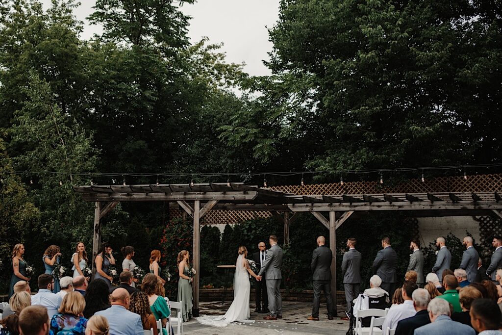 During their wedding ceremony at their venue, The Ivy House, a bride and groom stand holding hands with their wedding parties on either side of them and their guests seated watching the outdoor ceremony