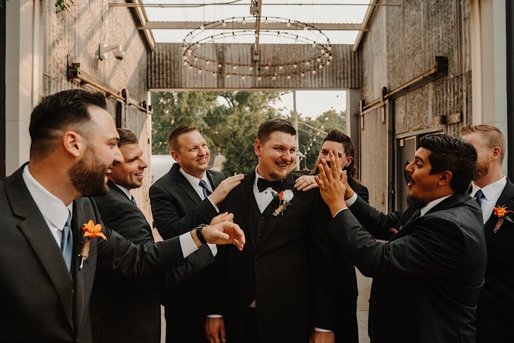 A groom smiles while standing in the middle of his 6 groomsmen under an outdoor chandelier as they all put their hands on his shoulders and hype him up