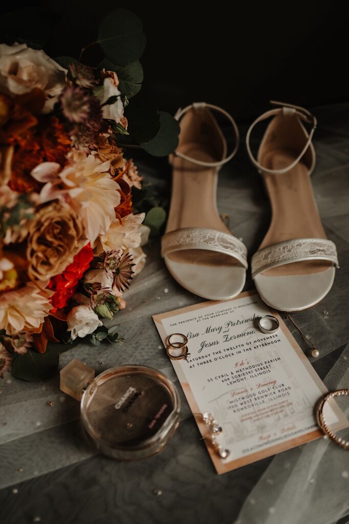 A wedding flat lay consisting of a wedding invite, rings, garter, bracelet, perfume, a veil, shoes, and flowers