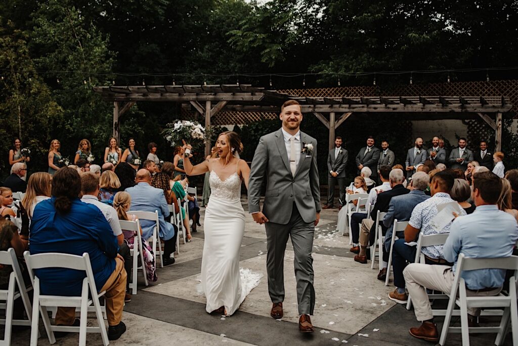 A bride and groom hold hands and walk down the aisle together after their outdoor wedding ceremony at their venue The Ivy House in Milwaukee, with guests cheering for them on either side