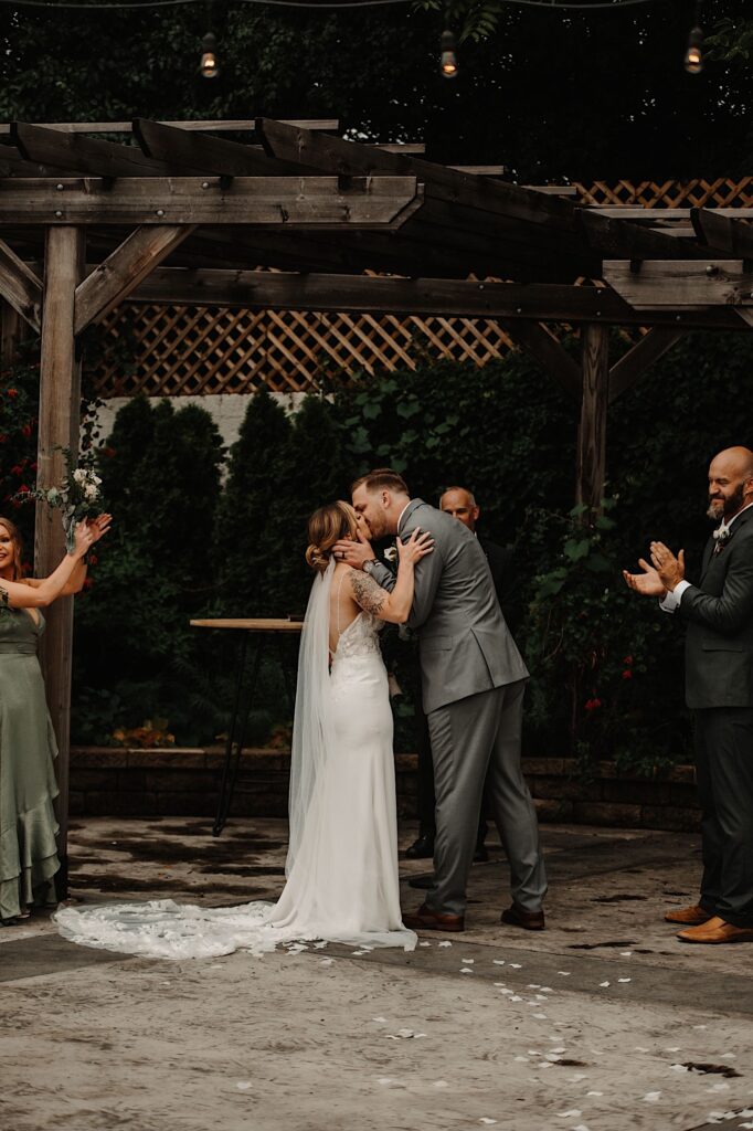 A bride and groom kiss one another during their outdoor wedding ceremony as their wedding party members and officiant look on and clap for them