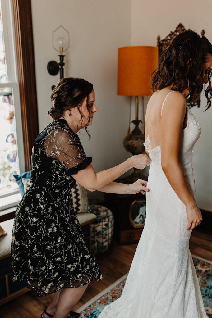 A bride stands in a room while her mother stands behind her and helps zip up her wedding dress