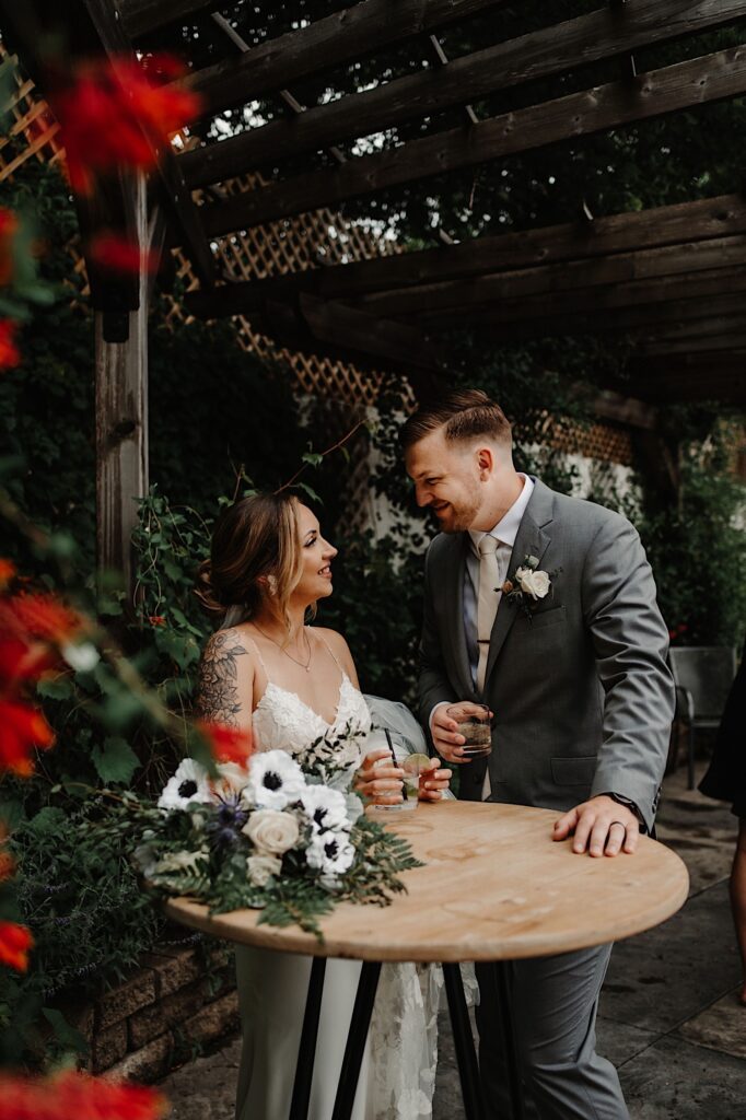 A bride and groom smile at one another while standing at a round table with drinks in their hand, they are outdoors with greenery all around them and the bride's bouquet on the table in front of them