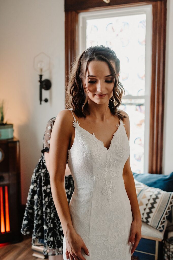 A bride in her wedding dress smiles while looking down standing in front of a window as her mother behind her helps zip up her dress