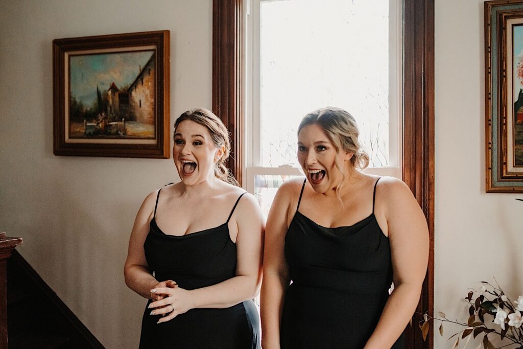 Two bridesmaids exclaim with excitement while standing in front of a window when they see the bride in her wedding dress for the first time