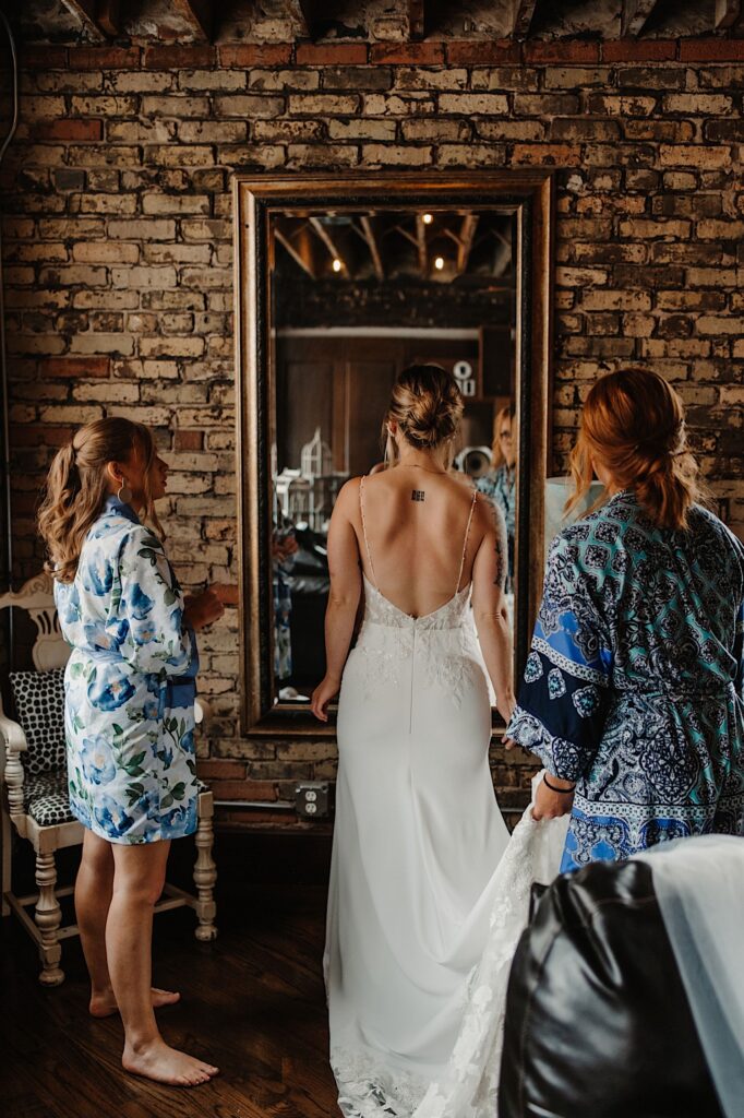 A bride in her wedding dress stands facing away from the camera looking at a mirror on a brick wall, she has a bridesmaid on either side of her helping her get ready for her wedding day