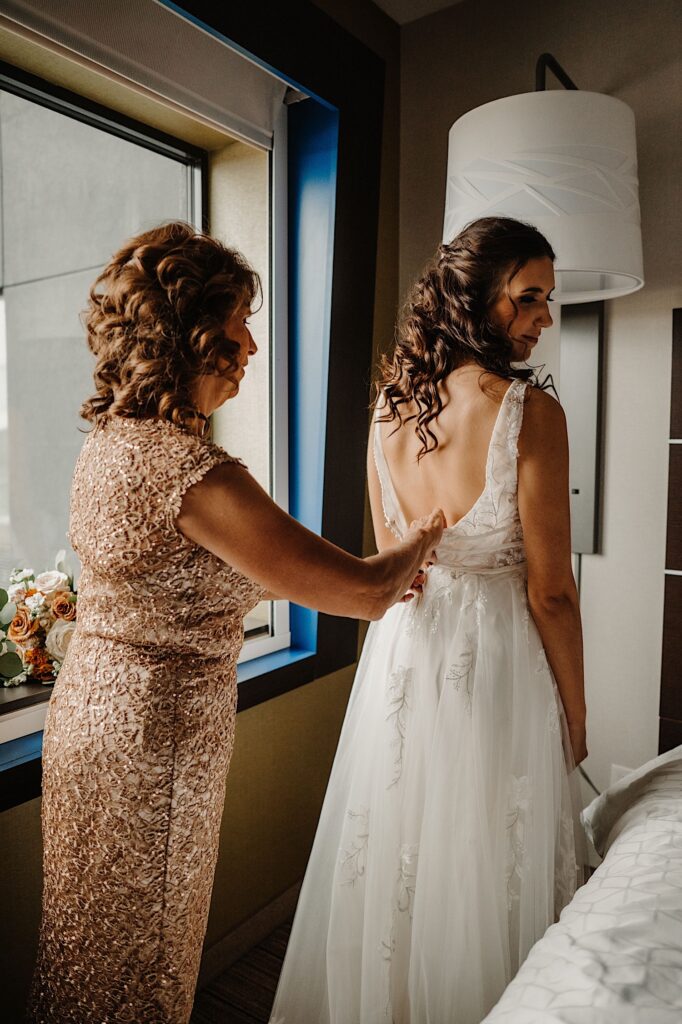 A bride stands with her back facing the camera with a window to her left as her mother helps button up the back of her wedding dress