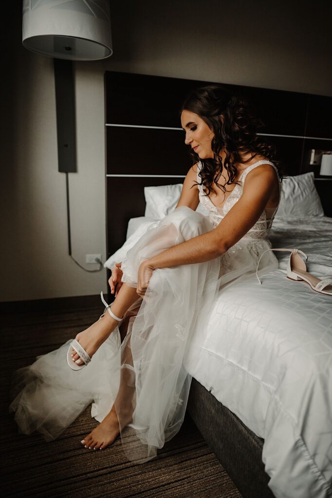 A bride sits on a bed while in her wedding dress and puts on her wedding shoes