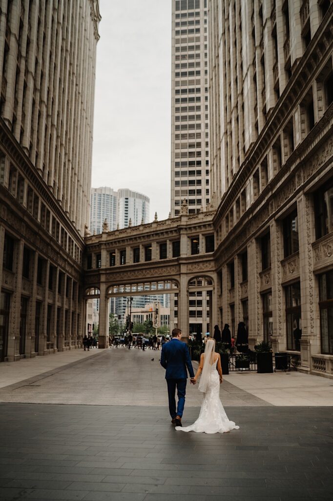 A bride and groom walk hand in hand next to a building in Chicago while facing away from the camera