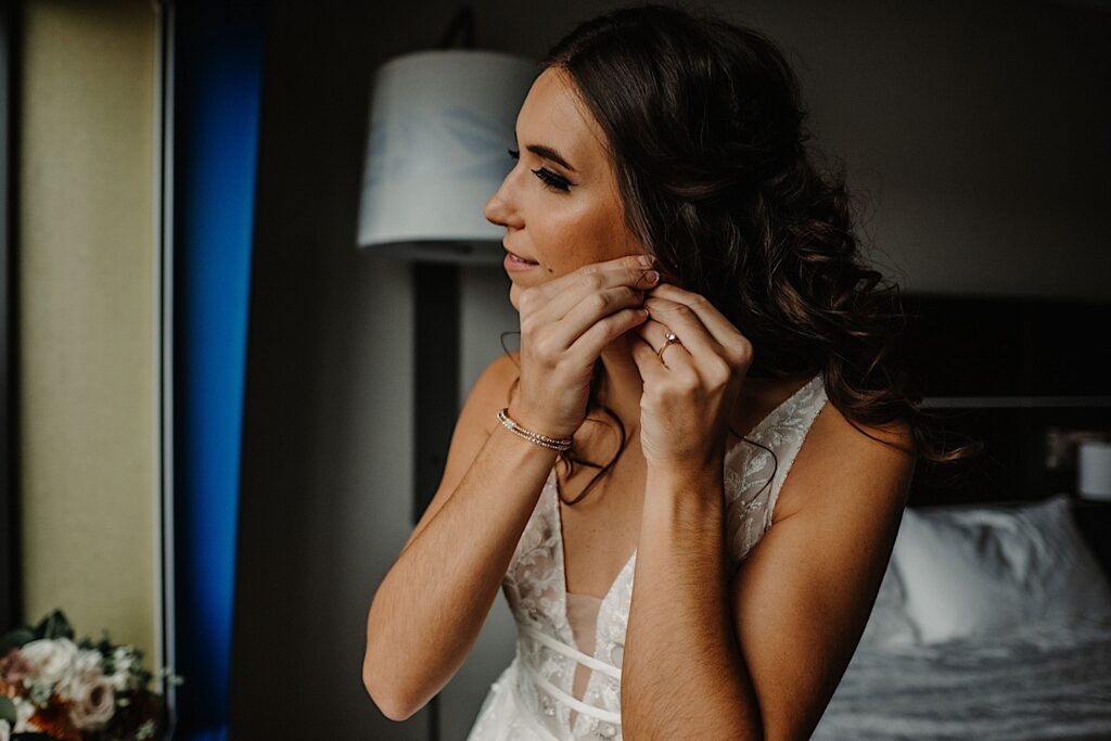 A bride sitting on a hotel bed in her wedding dress puts an earring on
