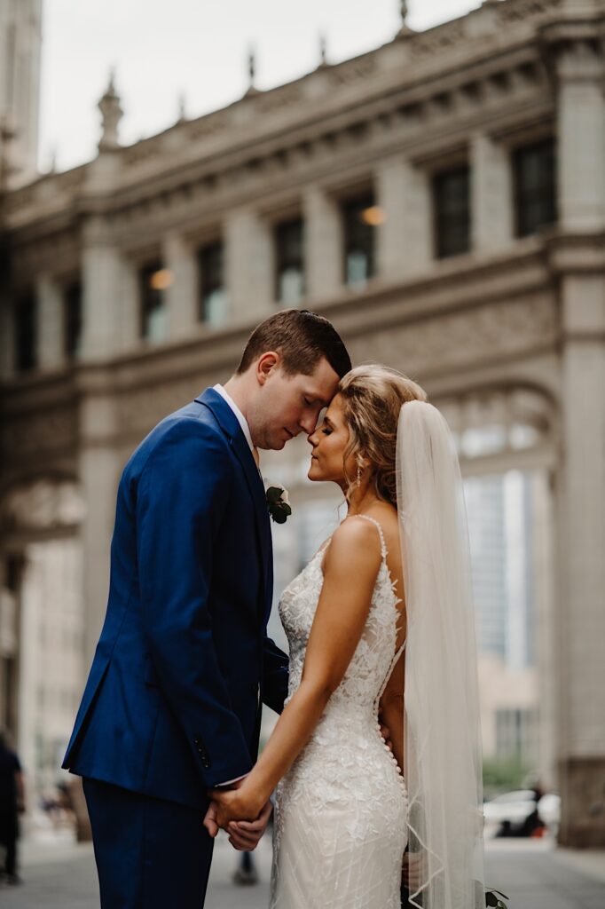 A bride and groom hold hands and face one another while touching foreheads with their eyes closed in front of a building in Chicago