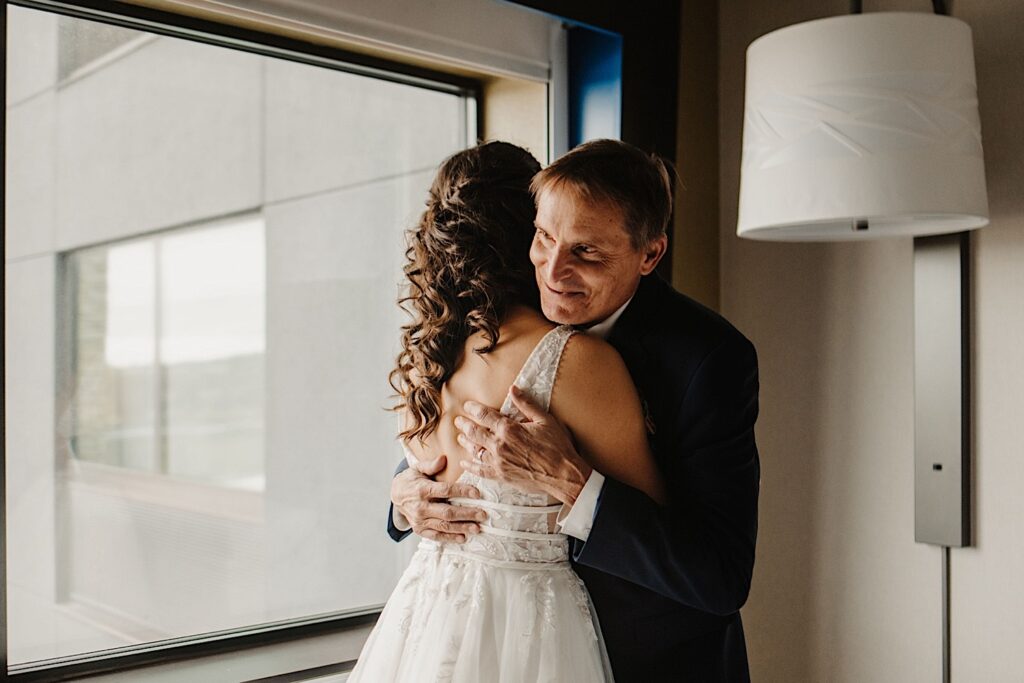 A bride hugs her father while in her wedding dress as he smiles, they're in a hotel room as the bride has just gotten ready for her wedding day