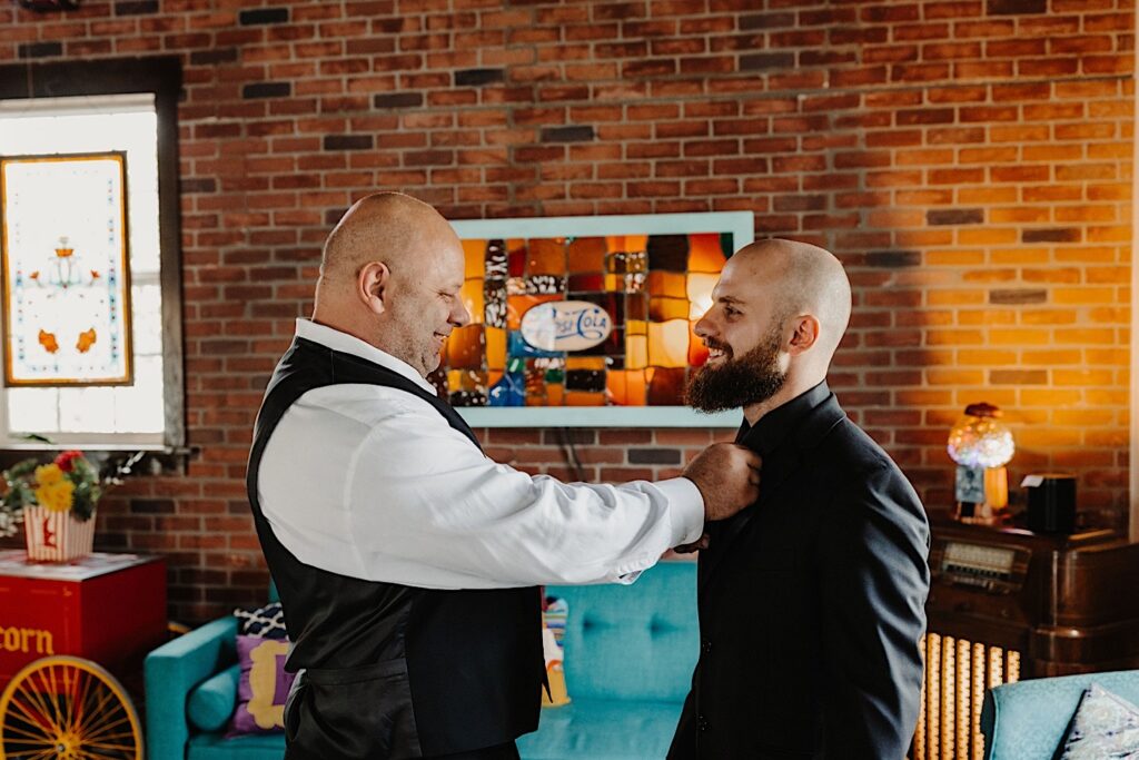 A groom smiles while standing in a brick room while his father smiles as he adjusts his tie