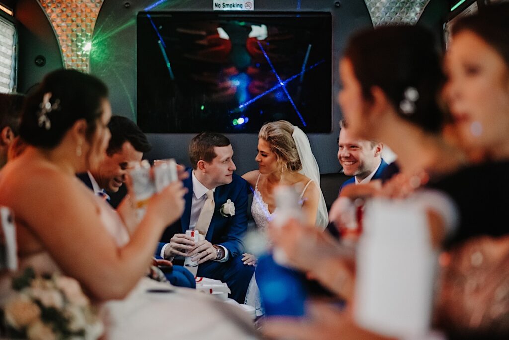 A bride and groom sit next to one another and smile at each other while on a party bus with their wedding parties sitting on either side of them