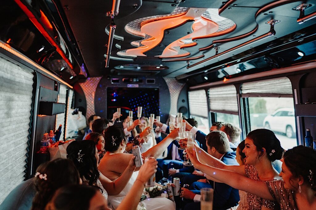 A wedding party raise their drinks along with the bride and groom while they all ride along on a party bus together