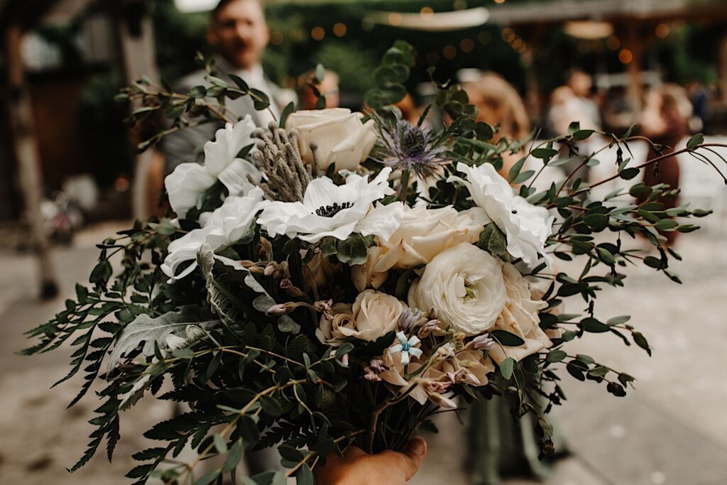 A bouquet of flowers for a wedding is being held out in front of the camera for a detail photo, the groom can be seen in the background but is out of focus