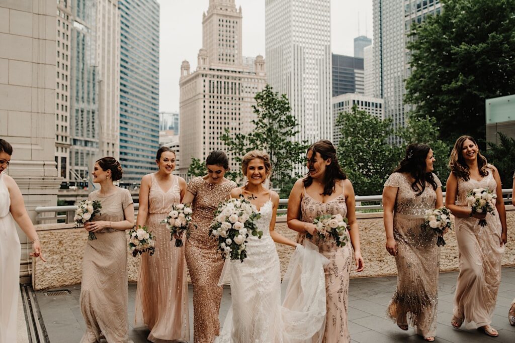 A bride walks towards the camera and smiles at it while in the middle of her 8 bridesmaids who are all walking and smiling as well with the Chicago skyline behind them